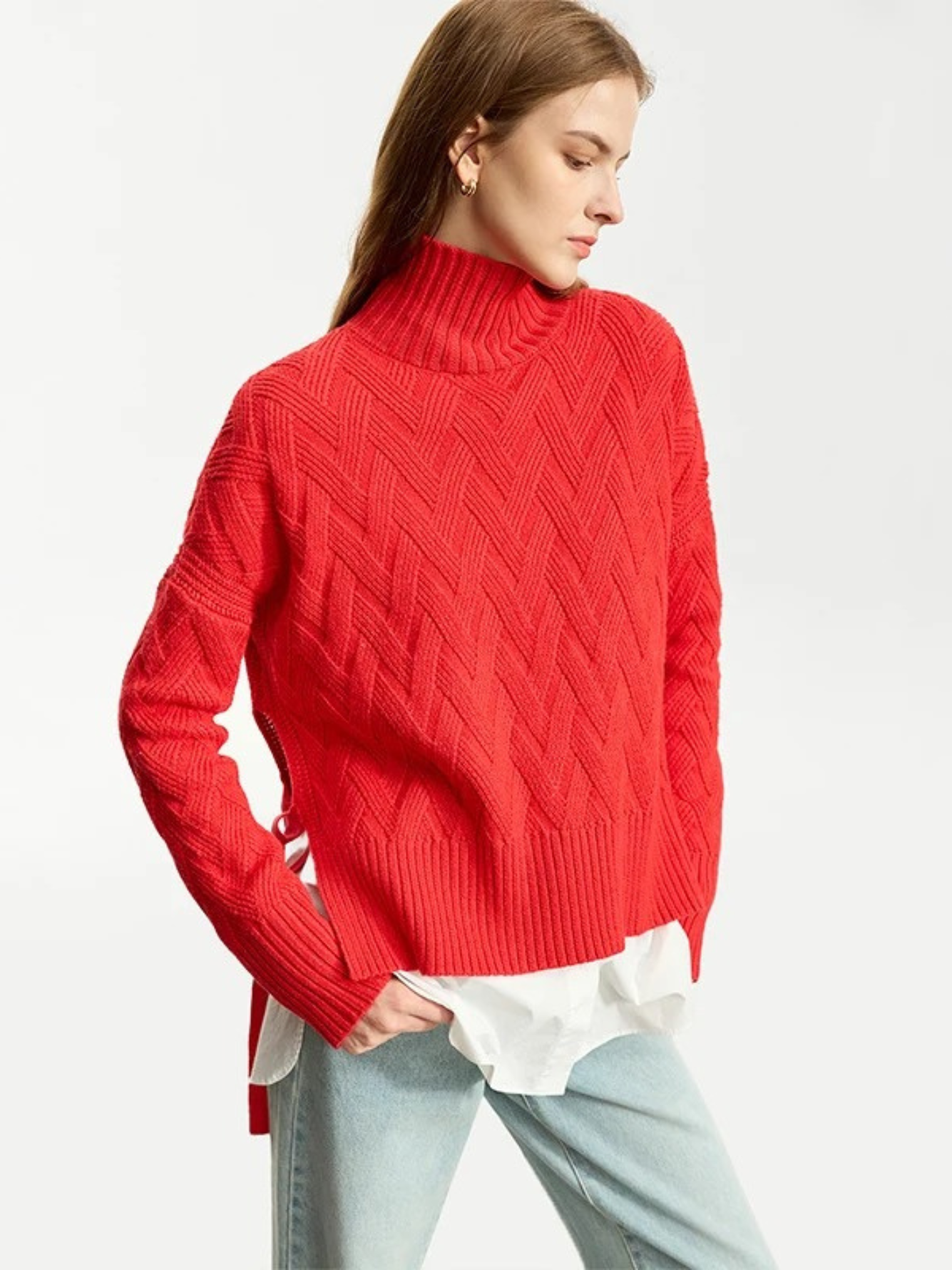 A woman wearing a turtle neck knit sweater with a tie design on the side in a bold red color with a white shirt underneath and a pair of brown light washed jeans.