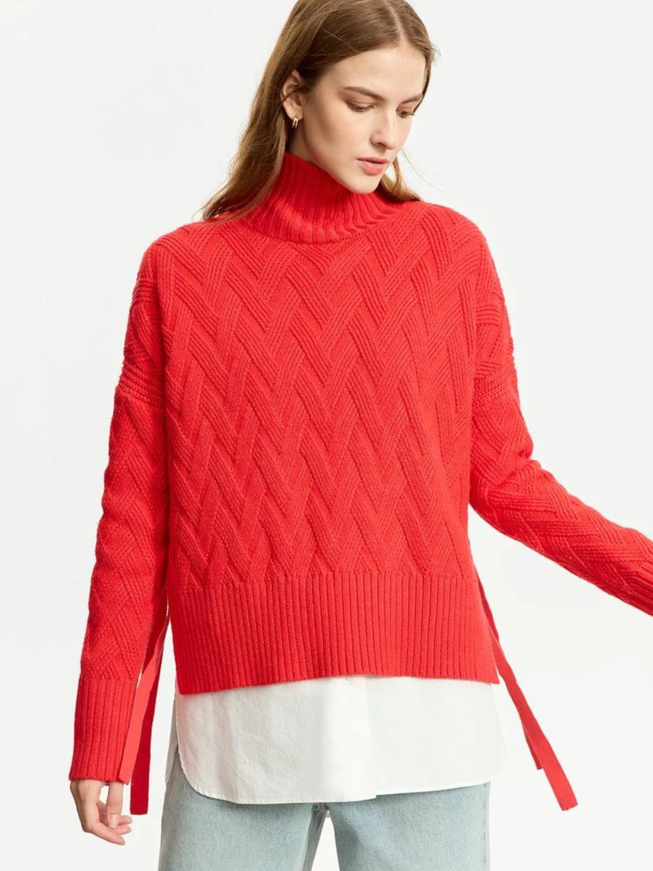A woman wearing a turtle neck knit sweater with a tie design on the side in a bold red color with a white shirt underneath and a pair of brown light washed jeans.
