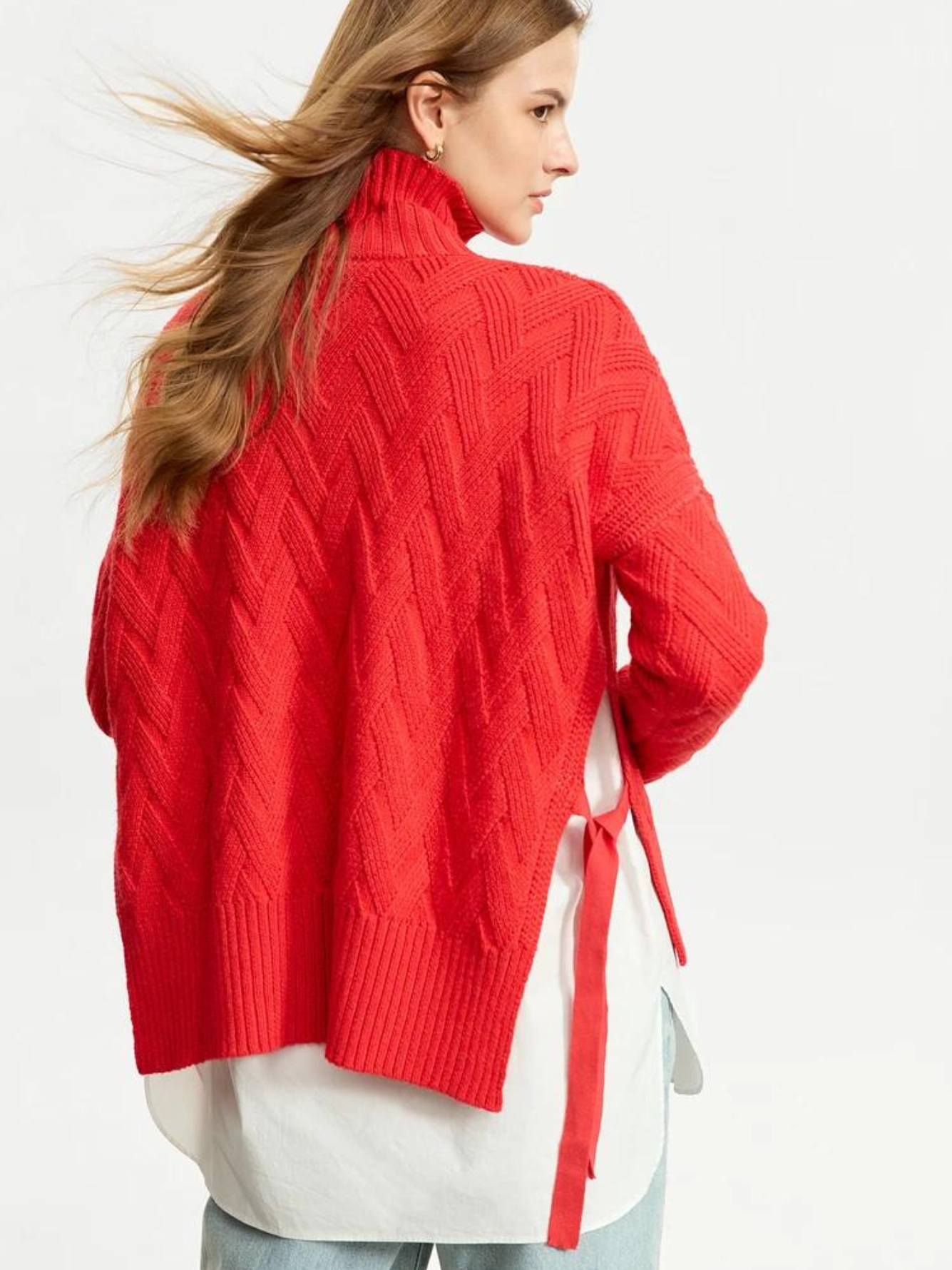 A woman with her back turned wearing a turtle neck knit sweater with a tie design on the side in a bold red color with a white shirt underneath and a pair of brown light washed jeans.