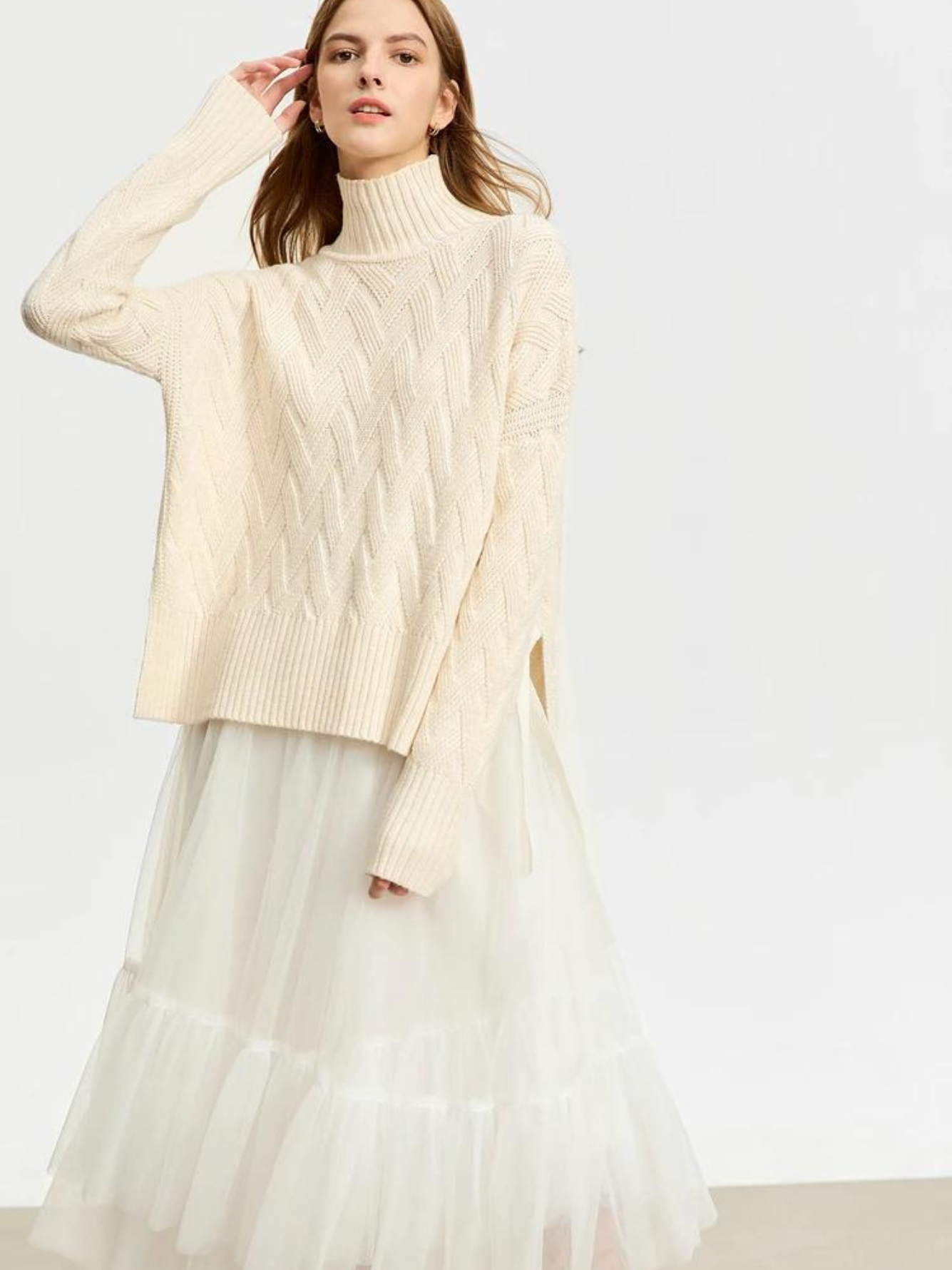 A woman wearing a turtle neck knit sweater with a tie design on the side in a cream color and a white tulle skirt.