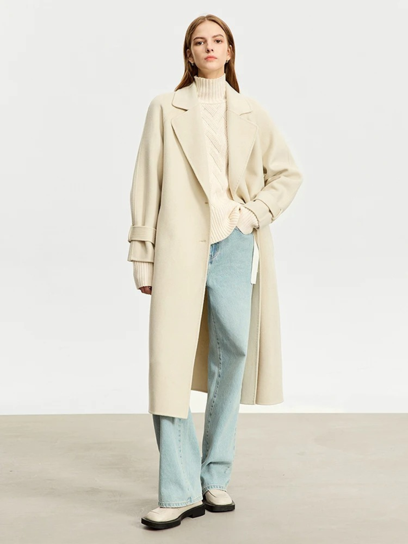 A woman wearing a turtle neck knit sweater with a tie design on the side in a cream color and a pair of denim jeans and an oversized trench coat.