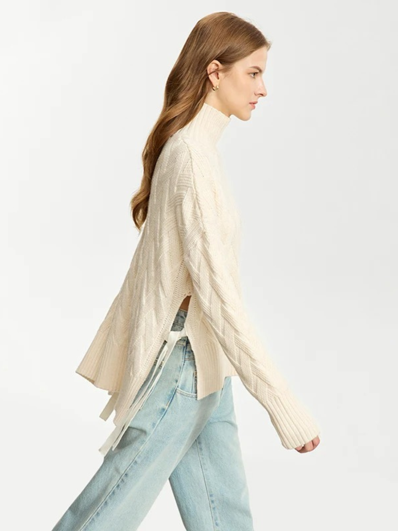 Profile of a woman wearing a turtle neck knit sweater with a tie design on the side in a cream color and a pair of washed jeans.