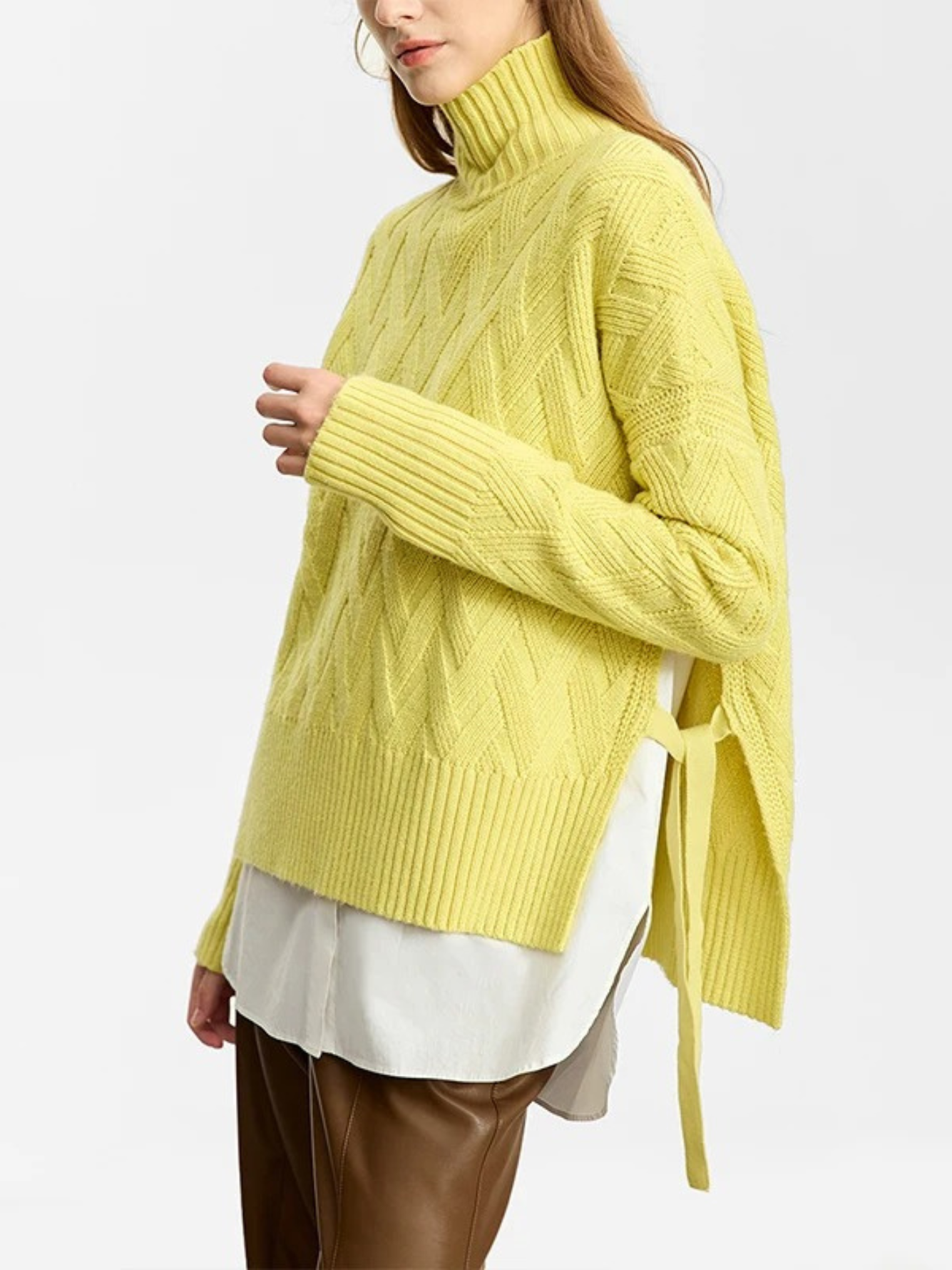A woman wearing a turtle neck knit sweater with a tie design on the side in a lime green color with a white shirt underneath and a pair of brown leather pants