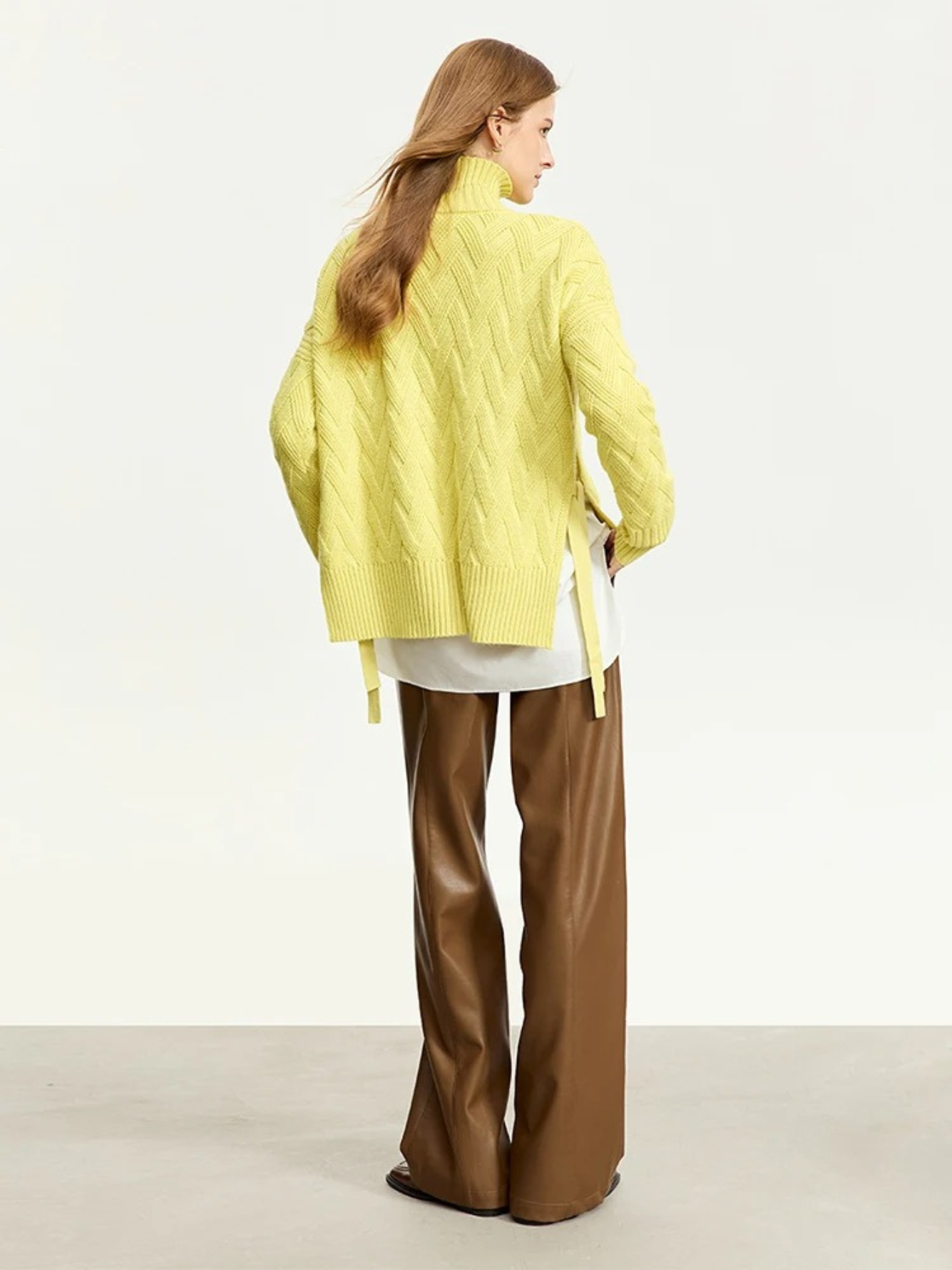 A woman with her back turned wearing a turtle neck knit sweater with a tie design on the side in a lime green color with a white shirt underneath and a pair of brown leather pants