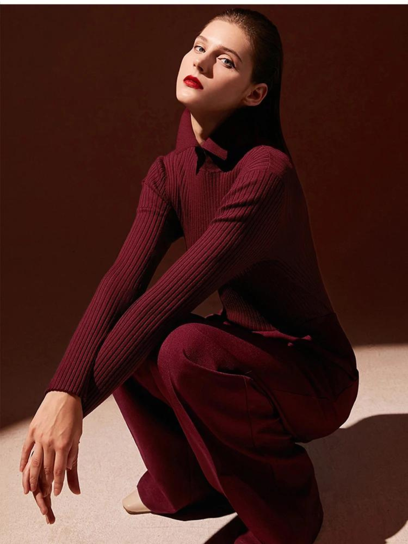 A woman wearing a ribbed sweater with a classic polo collar in red with a pair of red trousers against a dark background.
