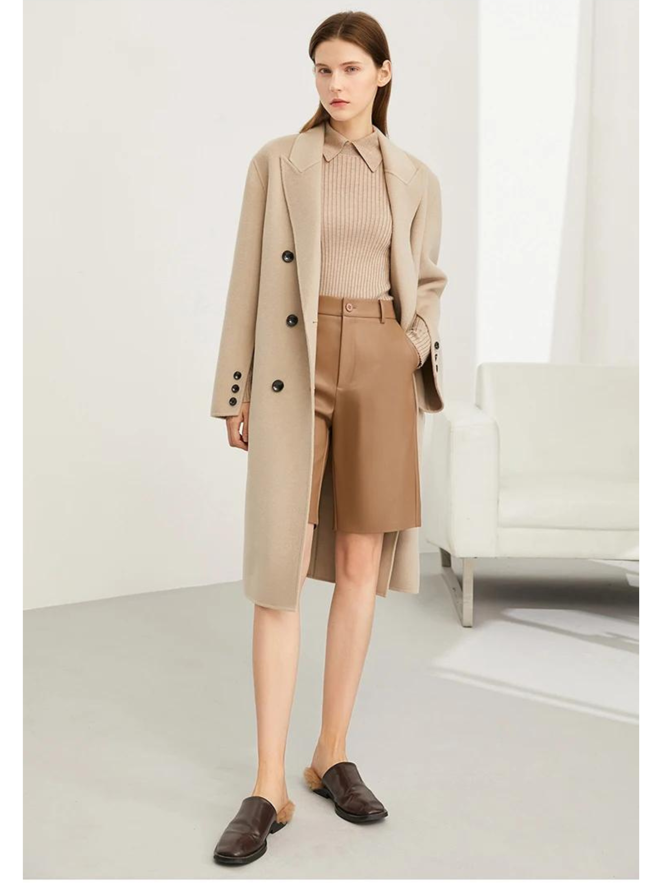 A woman wearing a ribbed sweater with a classic polo collar in khaki with a pair of brown leather shorts and a khaki trench coat.