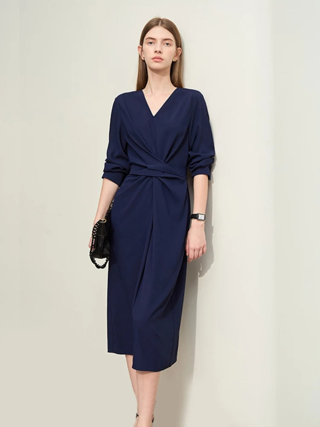 A stylish woman wearing a navy blue, long sleeved, midi dress with a ruching and tie design to cinch the waist,  and a slit at the back for ease of movement. The sleeves are rolled up to her elbows for a more casual look.