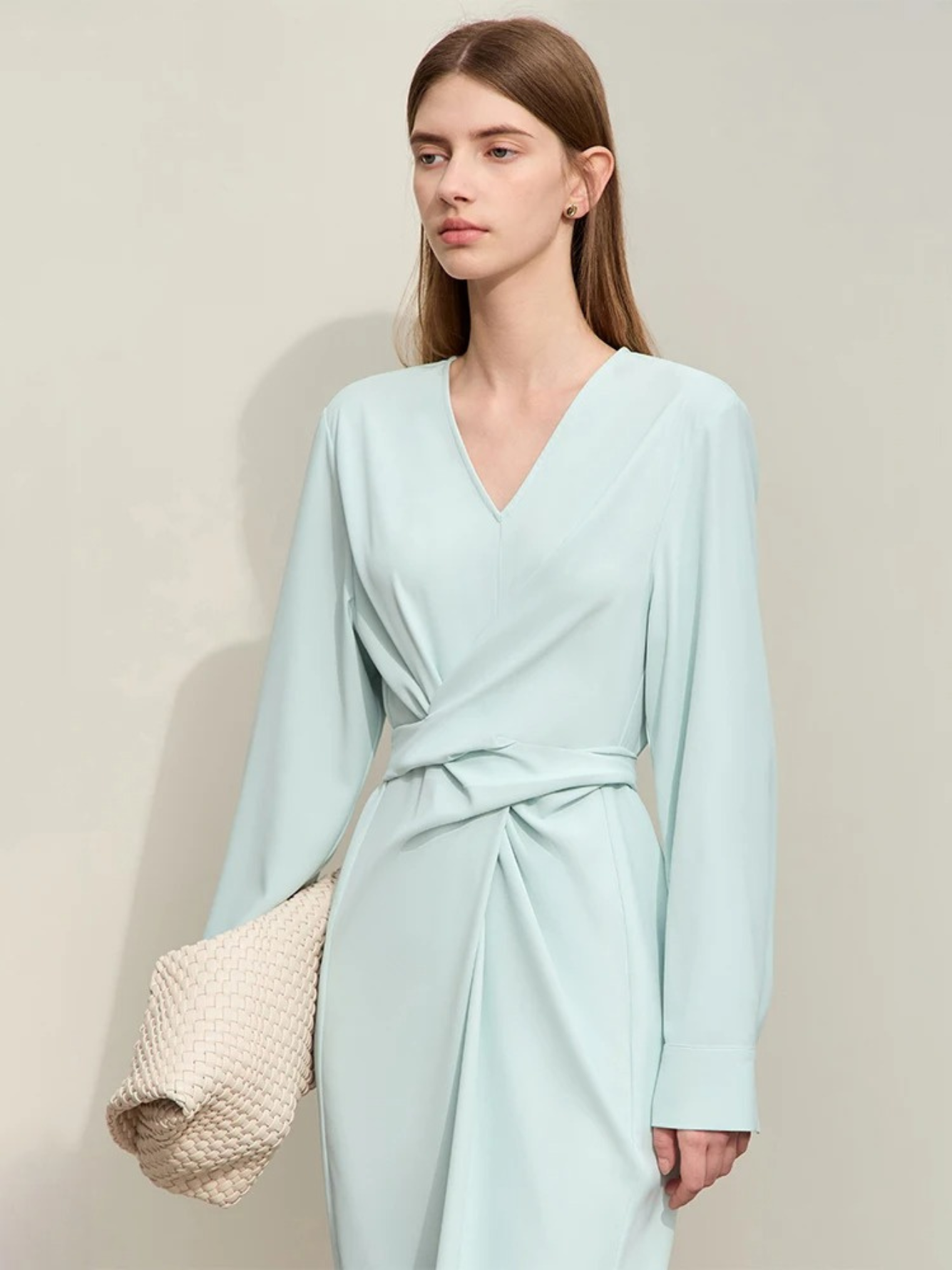A stylish woman wearing a powder blue, long sleeved, midi dress with a ruching and tie design to cinch the waist,  and a slit at the back for ease of movement. She paired it with a patterned white bag to complete the look.