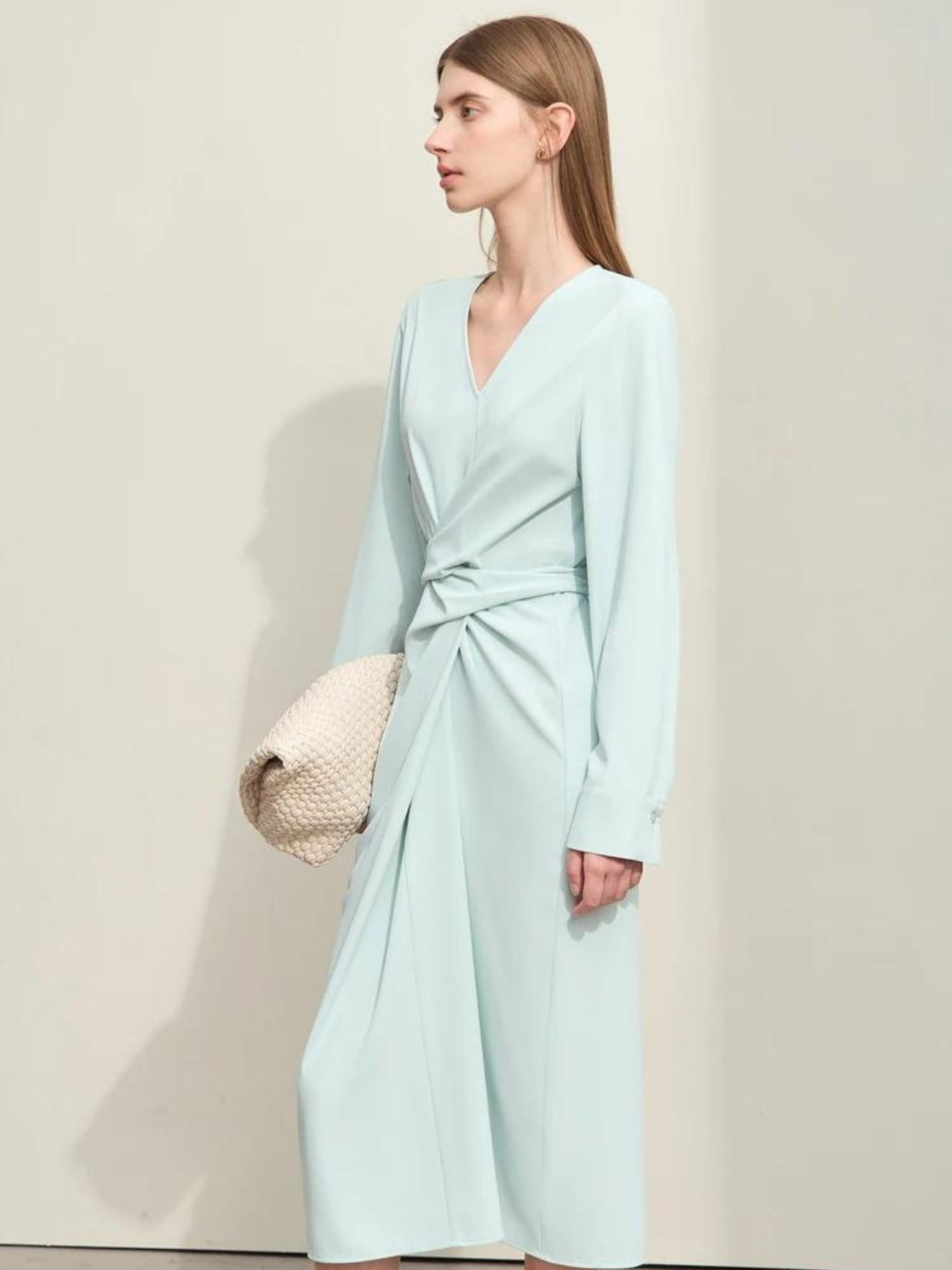 A stylish woman wearing a powder blue, long sleeved, midi dress with a ruching and tie design to cinch the waist,  and a slit at the back for ease of movement. She paired it with a patterned white bag to complete the look