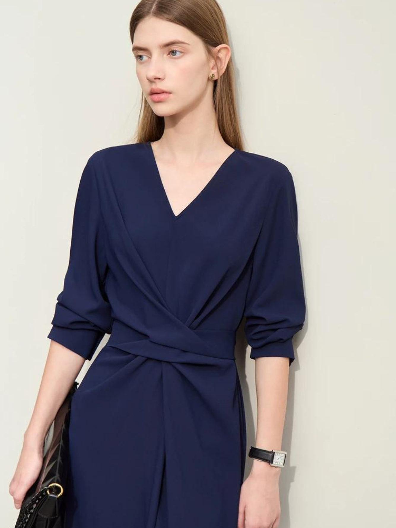 A stylish woman wearing a navy blue, long sleeved, midi dress with a ruching and tie design to cinch the waist,  and a slit at the back for ease of movement. The sleeves are rolled up to her elbows for a more casual look.