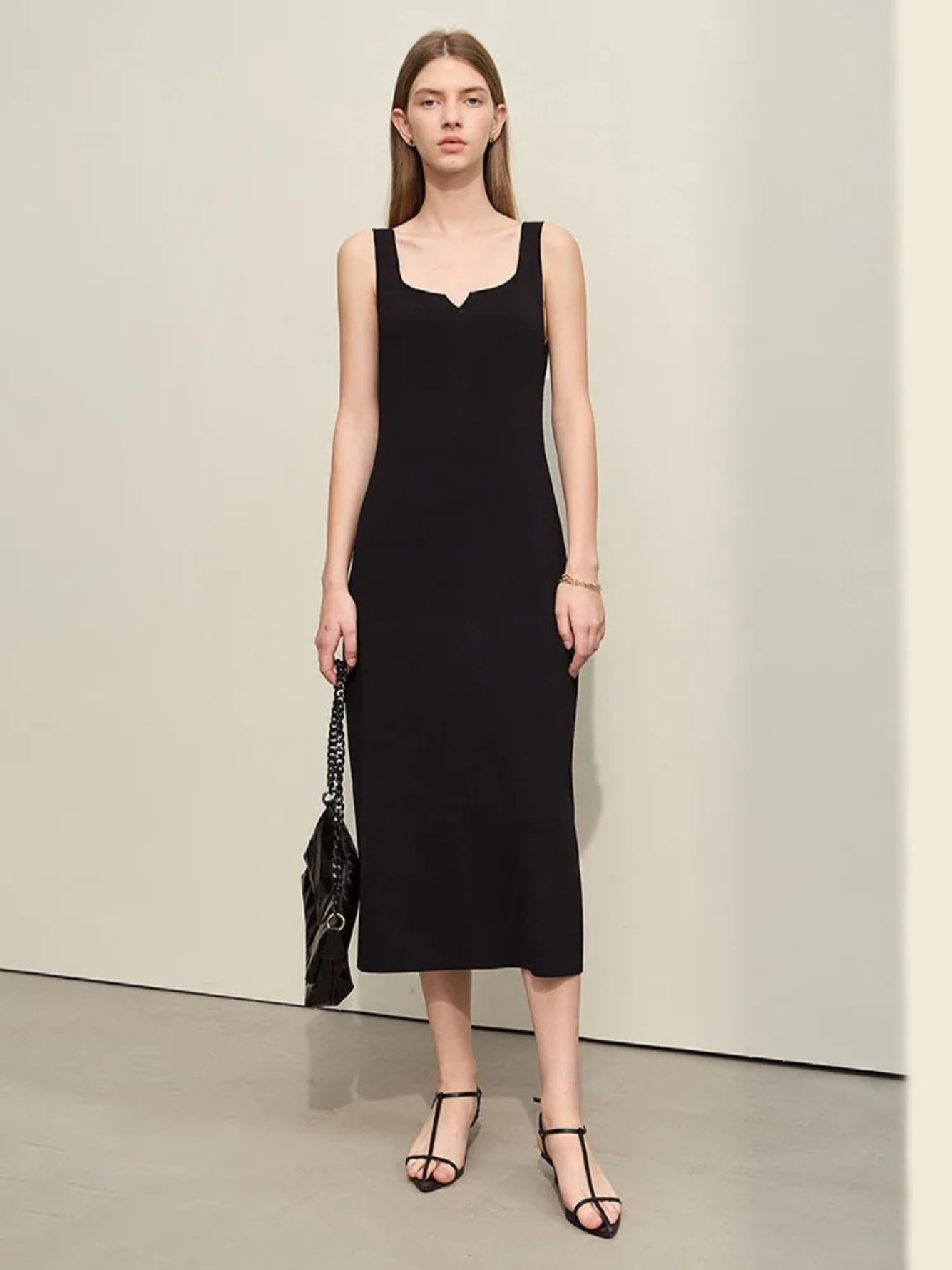 A woman wearing a simple, minimalist, sleeveless midi dresses with a v-cut out in the neckline in black. She has on black strappy heels and a black bag to finish the look.