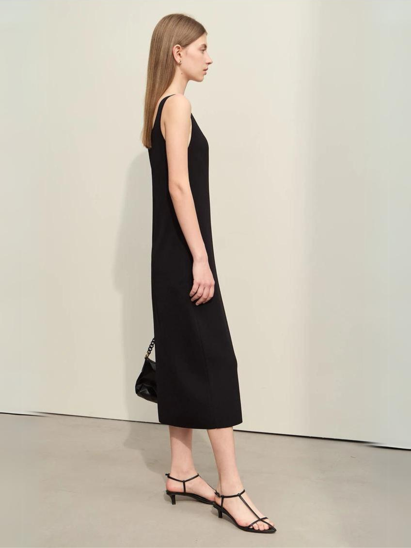 Profile view of a woman wearing a plain, minimalist, sleeveless midi dresses with a v-cut out in the neckline in black. She has on black strappy heels and a black bag to complete the look.
