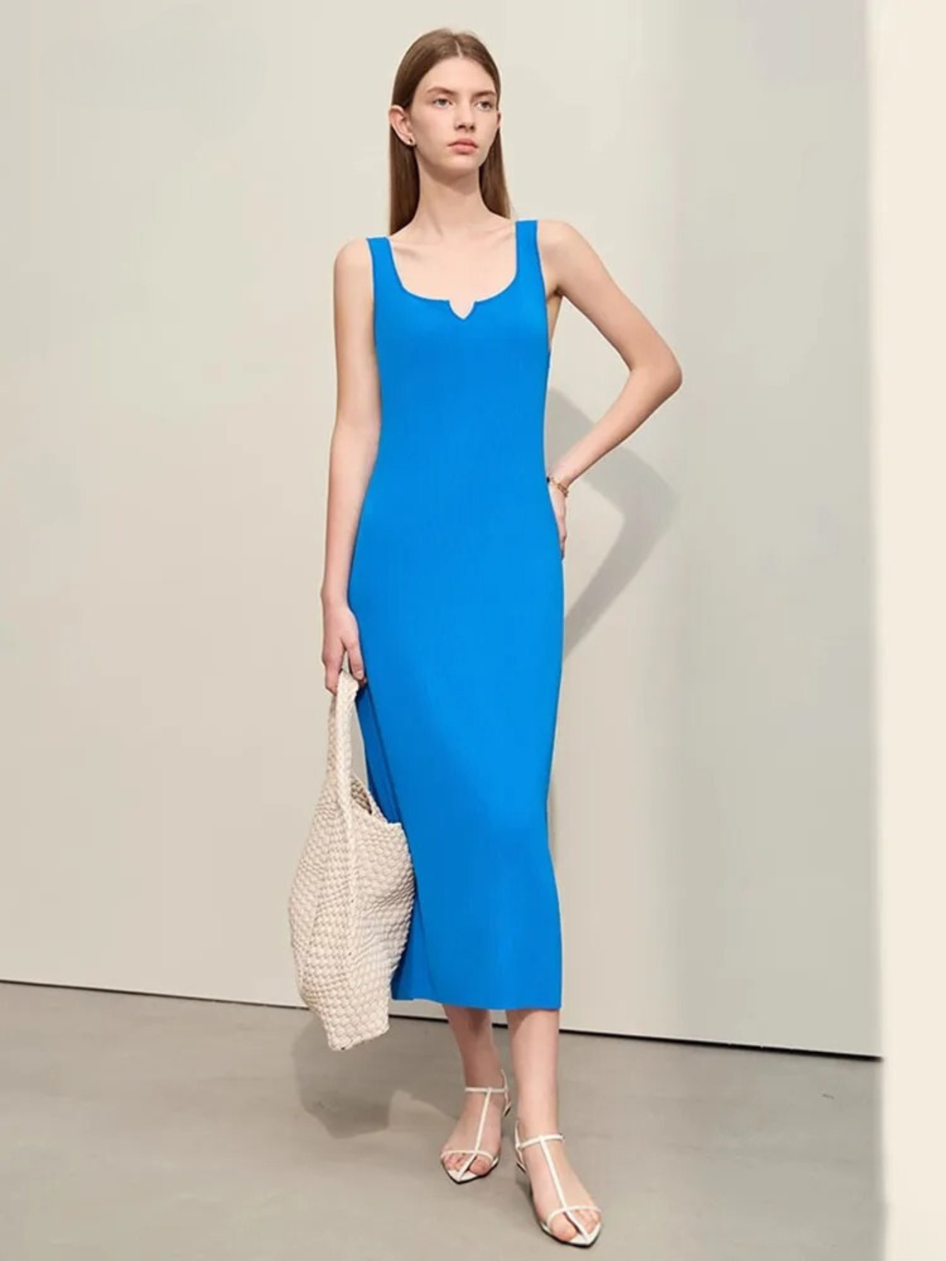 A woman wearing a plain, minimalist, sleeveless midi dresses with a v-cut out in the neckline in blue. She has on a pair of white strappy heels and a white bag to complete the look.