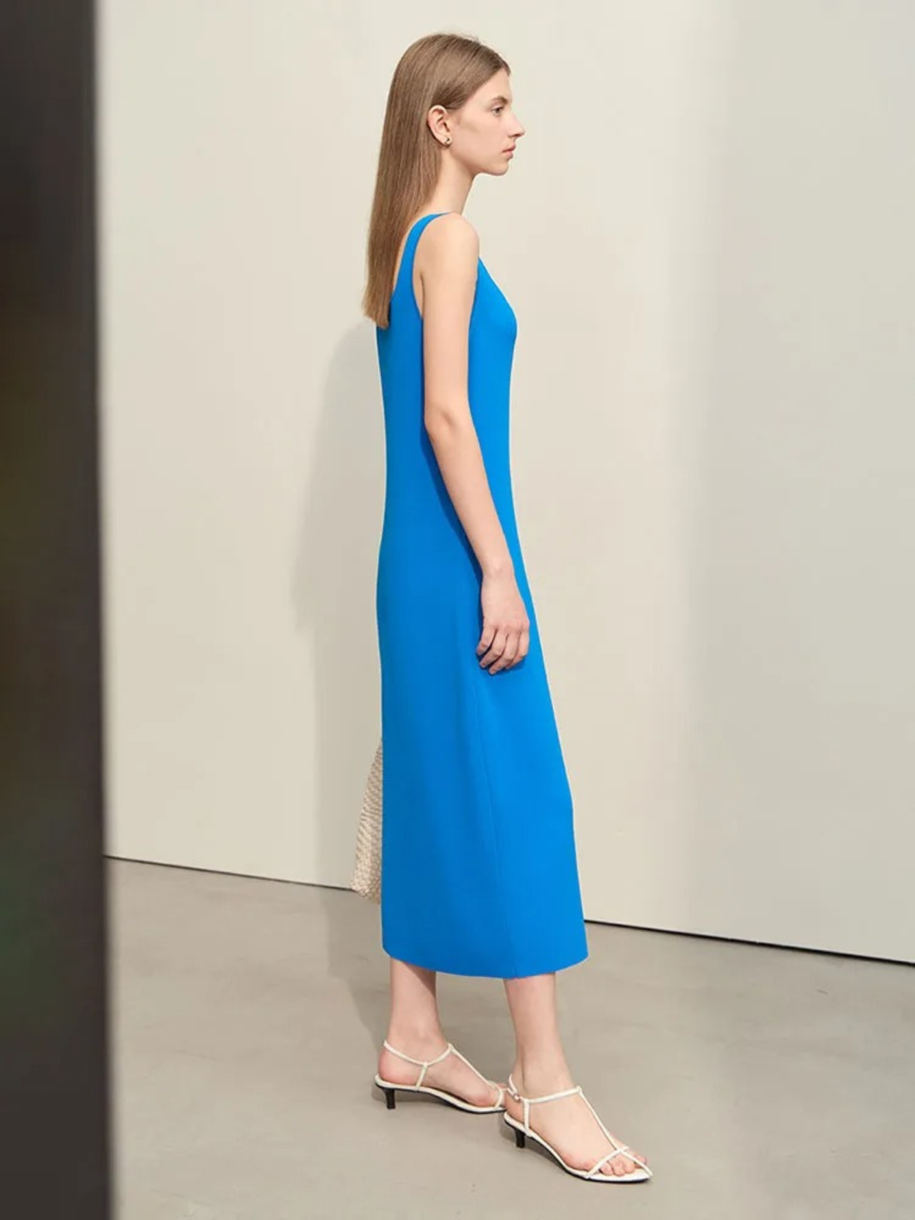 Profile view of A woman wearing a plain, minimalist, sleeveless midi dresses with a v-cut out in the neckline in blue. She has on a pair of white strappy heels and a white bag to complete the look.