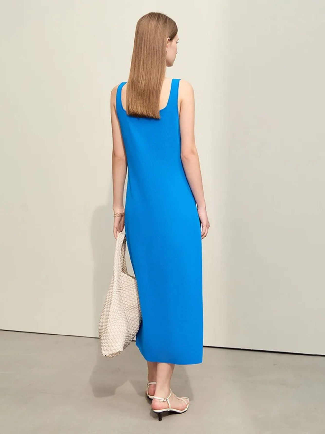 A woman turned away wearing a plain, minimalist, sleeveless midi dresses with a v-cut out in the neckline in blue. She has on a pair of white strappy heels and a white bag to complete the look.
