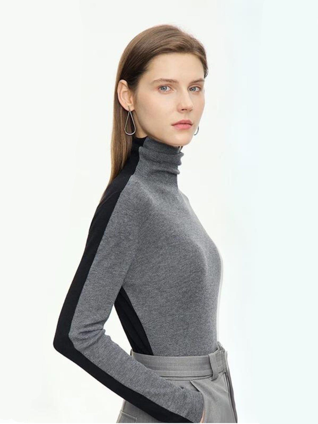 A woman wearing a mock neck, dual toned sweater in gray and black with a pair of gray belted high waisted pants with a belt detail with her hands in the pant pocket.