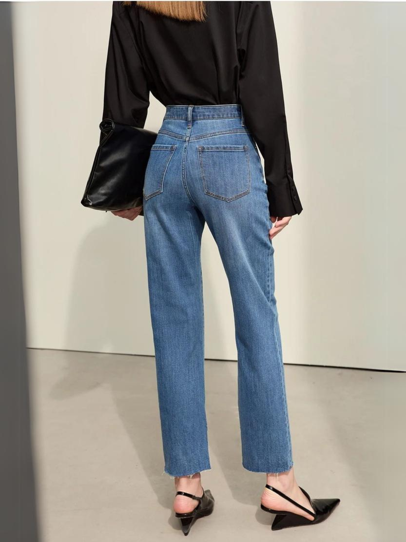 The backside of a woman wearing a classic black long sleeve shirt with a pair of denim blue, pencil cut jeans and a pair of heels.